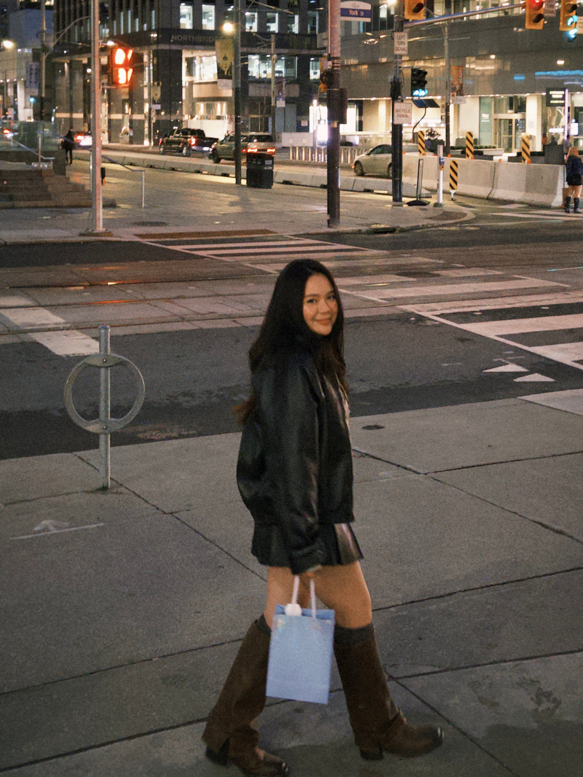 Woman in a leather jacket and knee-high boots standing on a city street at night.